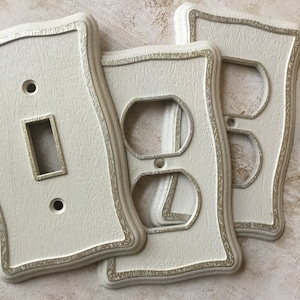 Op de afbeelding: Drie crèmekleurige kunststof licht schakelaar en stopcontact afdekplaten met een decoratieve zilveren rand. De afdekplaten hebben een golvend, sierlijk ontwerp. Vier schroeven zijn inbegrepen.