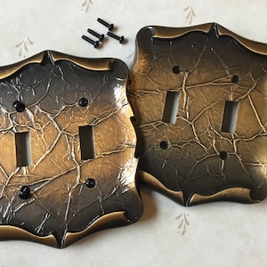 May include: Two decorative light switch covers with a textured, metallic finish. The covers have a dark bronze and gold colour scheme with a unique, crinkled appearance. Small black screws are included. The covers have a double switch design.