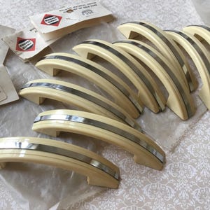 Peut inclure: Ensemble de poignées de meubles vintage crème avec une bande argentée. Plusieurs poignées sont disposées ensemble, certaines encore dans leur emballage d'origine. L'emballage porte l'inscription "WINNIPEG Supply & Fuel".