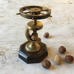 May include: A brass nutcracker with a wooden base and a wheel-shaped top. The nutcracker is holding a nut in its jaws. There are several nuts scattered around the base.