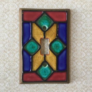 Puede incluir: Una cubierta decorativa para interruptor de luz con un diseño de vidriera. La cubierta tiene forma rectangular con un borde marrón. El diseño de vidriera incluye secciones rojas, azules, verdes y amarillas, con un interruptor plateado en el centro.