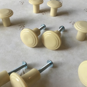 Op de afbeelding: Een verzameling crèmekleurige kastknoppen met een paddenstoelvormig ontwerp. De knoppen hebben een afgeronde bovenkant met concentrische cirkels en zijn gecombineerd met zilveren schroeven. De knoppen zijn waarschijnlijk voor meubels of kasten.