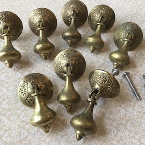 May include: A collection of antique brass drawer pulls with ornate, embossed detailing. Each pull features a round backplate and a teardrop-shaped handle. Several screws are included, suggesting a hardware set.
