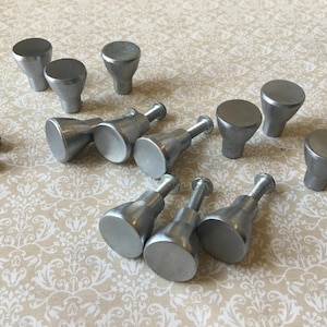 May include: A selection of silver cabinet knobs and screws. The knobs have a rounded top and a conical base. Some knobs are attached to screws. The items are arranged on a patterned surface.