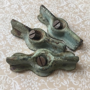 May include: Three antique, weathered metal hardware pieces with a distressed, turquoise-green patina. Each piece features a central screw and a unique, stylized shape, suggesting a vintage or industrial aesthetic. The hardware is arranged on a cream-colored, patterned surface.