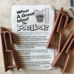 Puede incluir: Tres ganchos para macetas de color cobre con un anuncio. Los ganchos están diseñados para colgar macetas de terracota. El texto del anuncio dice "What a Great Idea! The Pot Hook". Incluye tornillos y tacos.