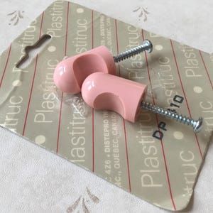 May include: Two pink plastic cabinet knobs with silver screws. The knobs are on a packaging card with the text "Plasttruc" and "DP 10".