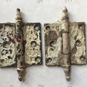 Puede incluir: Dos bisagras metálicas antiguas con diseños ornamentados y pintura blanca desgastada. Cada bisagra tiene un remate decorativo y múltiples orificios para tornillos. La pintura está desconchada, revelando el metal debajo, dándoles un aspecto vintage.