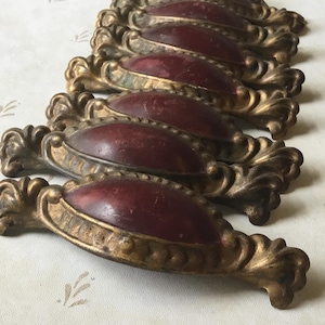 Op de afbeelding: Een verzameling antieke lade-knoppen. Elke knop heeft een bordeauxrode ovale kern, geplaatst in een goudkleurig, sierlijk frame. De knoppen hebben een gedetailleerd, vintage ontwerp.