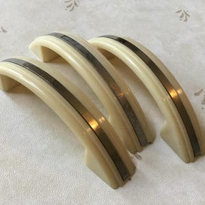 May include: Three vintage cabinet handles in a row. Each handle is a creamy off-white with a silver-coloured metallic stripe. The handles have a curved, arched design. They are made of a durable material.