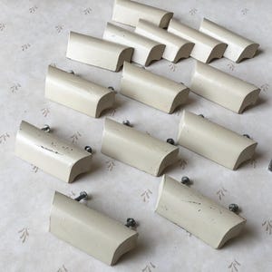 Puede incluir: Un juego de 12 pomos de armario de color blanco roto con forma rectangular redondeada. Los pomos tienen un borde ligeramente curvado y un solo agujero para tornillo.