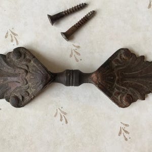 May include: An antique, ornate metal drawer pull with a symmetrical design featuring leaf-like details. Two rusty screws are positioned above the pull. The hardware is a dark, aged brown color.