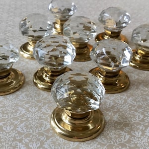 Peut inclure: Ensemble de neuf boutons de porte en cristal transparent avec des surfaces à facettes. Chaque bouton est monté sur une base ronde de couleur or. Les boutons sont disposés sur une surface blanche cassé à motifs.