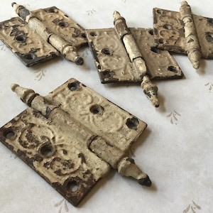 Puede incluir: Varias bisagras metálicas antiguas con un acabado desgastado en blanco roto. Las bisagras tienen un diseño ornamental y probablemente son para un armario o una puerta. El metal tiene un aspecto envejecido, con óxido y pintura descascarillada.