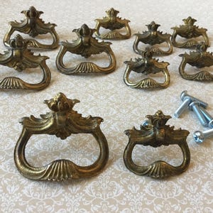 Peut inclure: Ensemble de poignées de tiroir antiques en laiton avec des motifs floraux et coquillages ornés. Les poignées sont disposées sur une surface à motifs, avec plusieurs vis. Ces pièces de quincaillerie décoratives sont idéales pour la restauration de meubles.