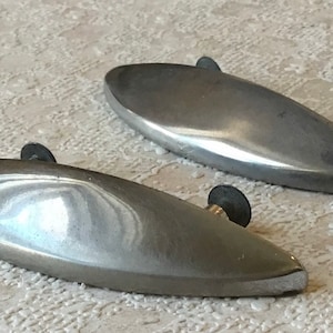 Puede incluir: Dos tiradores de cajones de forma ovalada de color plateado con pomos negros. Los tiradores son lisos y tienen una forma ligeramente curvada.