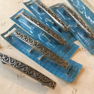 May include: Several silver-toned cabinet pulls with an ornate, scrolling design are displayed on blue packaging. The pulls are rectangular with curved handles and come with screws. The background is a light beige surface with a subtle floral pattern.
