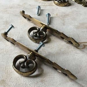 Peut inclure: Deux poignées de tiroir en laiton avec des volutes ornées et un motif de fleur de lys. Les poignées sont fixées à une base plate rectangulaire avec des vis.