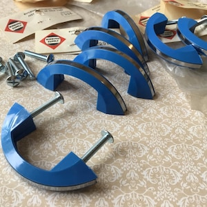 May include: A set of blue cabinet handles with silver detailing and screws. The handles, with their curved design, are arranged on a patterned surface. The image also shows several screws, suggesting a vintage style. The handles are approximately 10 cm long.