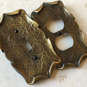 May include: Two decorative light switch and outlet covers with an antique finish. The covers have a textured, gold-toned appearance with black screws. The edges are detailed with a darker gold trim, enhancing the vintage look.