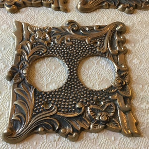 May include: A brass double outlet cover with an ornate, floral design. The cover has a textured surface and is in a vintage style.