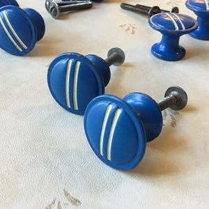 May include: A set of blue and white ceramic cabinet knobs with a simple design. The knobs have a round shape with two white stripes on a blue background.