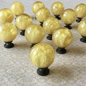 Peut inclure: Ensemble de boutons de meubles décoratifs. Chaque bouton est composé d'un dessus en verre sphérique transparent avec des inclusions dorées, monté sur une base sombre et ornée. Les boutons sont disposés sur une surface à motifs, d'un style vintage.