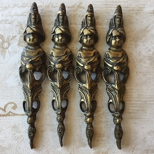 Könnte beinhalten: Satz von vier antiken, goldfarbenen, dekorativen Metallakzenten. Jeder Akzent zeigt ein detailliertes Design einer Figur mit einer Krone und verzierten Ranken.