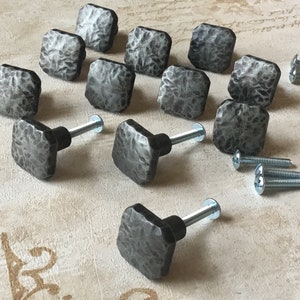 Può includere: Un set di 12 pomelli quadrati per armadi con finitura in metallo martellato. I pomelli sono di colore argento e hanno un aspetto rustico. Ogni pomello viene fornito con una vite per l'installazione.