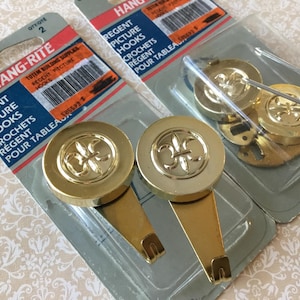 Può includere: Due confezioni di ganci per quadri Hang-Rite Regent. I ganci sono color oro con un design a giglio sulla testa rotonda. La confezione include il testo "Regent Picture Hooks" e "Pour Tableaux".