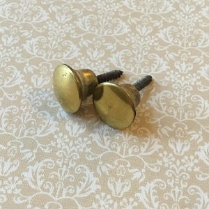 以下が含まれることがあります： ネジ付きの真鍮製ヴィンテージキャビネットノブ2個。ノブは丸みを帯びた平らなデザインで、暖かく金色の色合いです。 白い花柄のクリーム色の背景に配置されており、クラシックまたはアンティークな美しさを暗示しています。