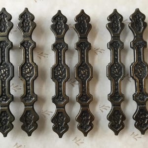 Puede incluir: Conjunto de seis tiradores de cajón de armario de latón antiguo con un diseño ornamentado. Cada tirador tiene un patrón decorativo intrincado y una forma puntiaguda ligeramente curvada.