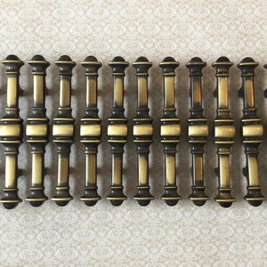 May include: A set of antique brass door pulls. Each pull has a dark bronze finish with gold detailing. The design includes a cylindrical body with decorative elements. The pulls are arranged in a row, suggesting they are ready for installation.