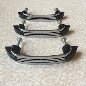 May include: Three cabinet handles with a black and silver design. The handles have a curved shape with three horizontal black stripes and are attached with silver screws. The ends of the handles are black.