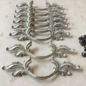 May include: A collection of decorative silver-coloured drawer pulls featuring a scrolling leaf design. The handles are arranged in rows, with a small pile of dark screws. These ornate pulls are suitable for furniture restoration.