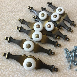 May include: A collection of antique-style drawer pulls with white ceramic knobs and bronze-toned metal bases. The pulls are arranged on a patterned surface, with a small pile of silver screws nearby.