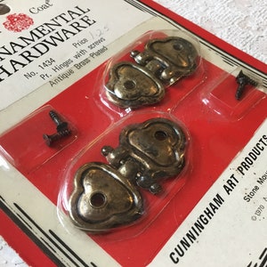 Puede incluir: Dos bisagras en forma de corazón de latón antiguo plateado con tornillos en una tarjeta de embalaje roja y blanca. La tarjeta dice "Royal Coat Ornamental Hardware No. 1434 Price 125 Pr. Hinges with screws Antique Brass Plated" y "CUNNINGHAM ART PRODUCTS, INC. Stone Mountain, Ga. 30083 ©1970 CAP, INC. 95190".