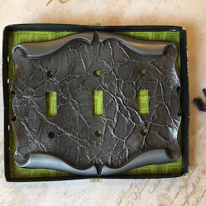 May include: A gray metal light switch plate with a textured, faux leather finish. The plate has three switch openings and a decorative, ornate border. The plate is in its original packaging, which is a black box with a green and white pattern.
