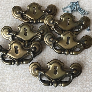 以下が含まれることがあります： 鍵穴のディテールと湾曲したハンドルを備えた、真鍮製のアンティーク引き出しプル6個セット。小さな銀色のネジが含まれています。 家具の修復やキャビネットにクラシックなタッチを加えるのに適した、ヴィンテージ感のあるハードウェアです。