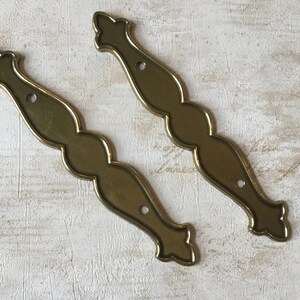 May include: Two ornate brass drawer pulls with a decorative, wavy design. Each pull has three holes for mounting.
