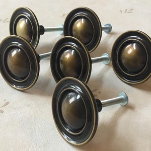 May include: Six antique brass cabinet knobs with a circular design and a raised center. The knobs have a dark brown finish and are attached to silver screws.