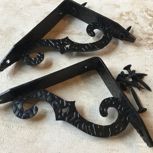 Puede incluir: Dos soportes de estante de metal negro con un diseño decorativo de desplazamiento. Los soportes están hechos de metal y tienen un acabado negro. Se muestran sobre una superficie beige claro.