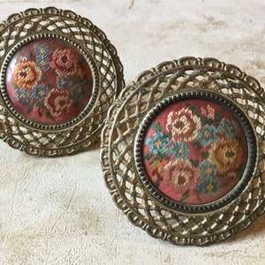 Könnte beinhalten: Zwei runde Vintage-Broschen aus Metall mit aufwendigen filigranen Rahmen. Jede Brosche hat ein kreisförmiges Mitteldesign mit einem floralen Kreuzstichmuster in Rot, Blau, Gelb und Grün. Das Metall hat ein antikes Finish.