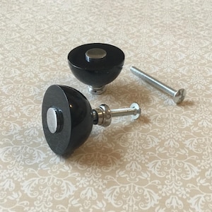 Op de afbeelding: Twee zwarte kastknoppen met zilveren accenten en een zilveren schroef. De knoppen hebben een afgeronde vorm met een platte bovenkant en een zilveren schijf in het midden. De schroef is lang en zilverkleurig. De knoppen liggen op een patroon.