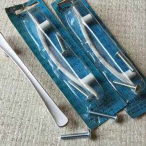 Può includere: Due confezioni di maniglie per ante di armadio in plastica bianca con un design curvo. Le confezioni sono etichettate "Amerock" e riportano il testo "Solid White Accent" e "Solid White Golden Accent".