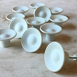 Pode incluir: Um grupo de 12 botões de armário de cerâmica branca com um centro elevado e uma superfície texturizada. Os botões estão dispostos em um padrão circular sobre uma superfície branca.