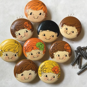 May include: A collection of nine round, hand-painted wooden knobs featuring smiling cartoon faces with various hairstyles and colors, including brown, orange, yellow, and black. A small pile of silver screws is included.
