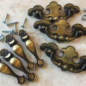 May include: Set of eight antique brass cabinet pulls. Four are ornate with a decorative scroll design and four are simple, elongated pulls. The pulls come with screws.
