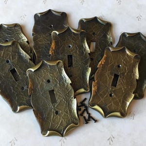May include: A selection of antique-style light switch covers with a textured, bronze finish. Each cover has a decorative, ornate edge and a rectangular opening for the switch. Small black screws are included.