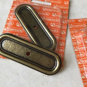 May include: Two gold-toned metal drawer pulls with a textured surface and a raised oval border. The pulls are packaged in clear plastic on a bright orange background with the word "Amerock" printed repeatedly.